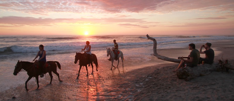 Horseback riders and surfers on Playa Carmen at sunset