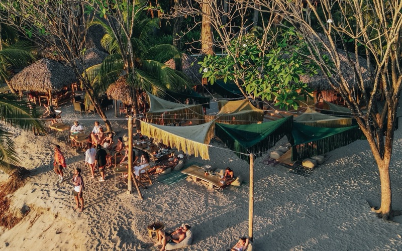 Aerial view of UMA's beach seating at sunset, with shade sails, low tables and thatched huts under the trees at Playa Cielo