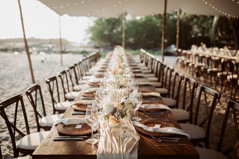 Long wooden dinner table set with wine glasses and flowers under a tent on the beach