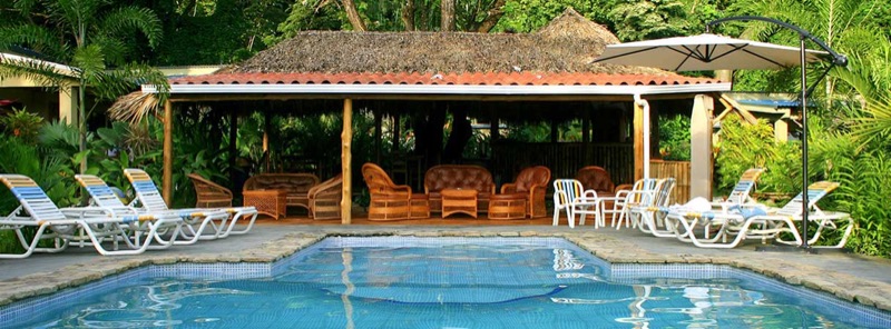 Pool in front of a thatched rancho with wicker sofas and white sun loungers, surrounded by tropical plants at Zula Inn